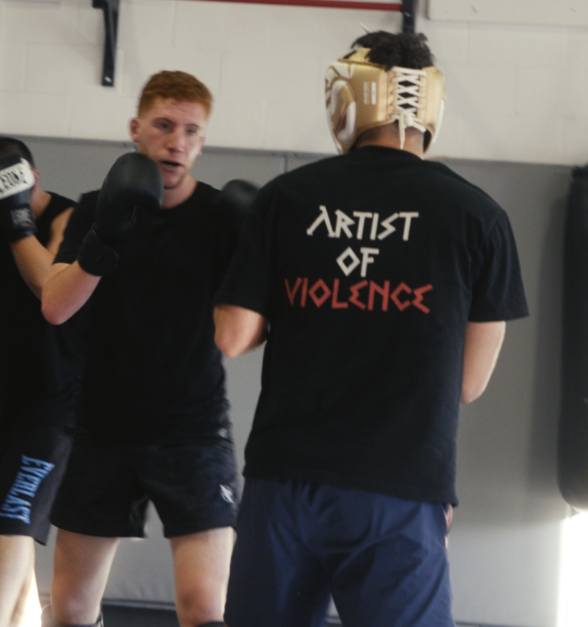 Two individuals in a boxing or martial arts training session with one wearing a 'Artist of Violence' shirt.